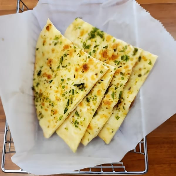 Four pieces of garlic naan bread, garnished with green herbs, served in a basket with white paper.