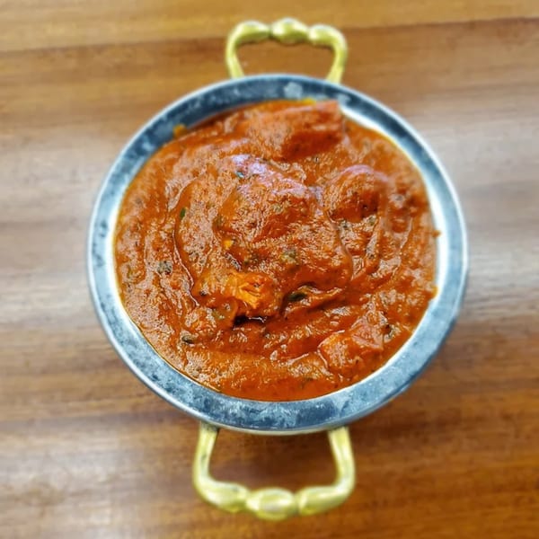 Indian curry, likely butter chicken, in a metal handi with brass handles on a wooden table.
